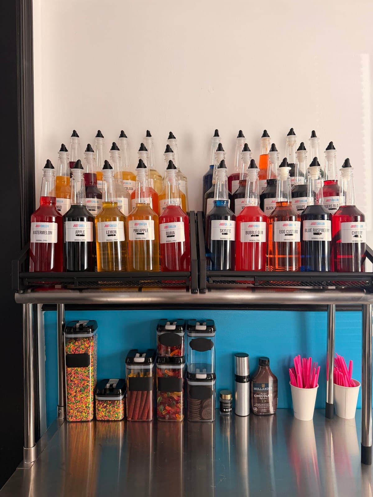 Rows of colorful flavor syrup bottles above a station of candy toppings and pink spoons at Just Chillin